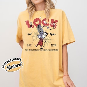 May include: Mustard-colored t-shirt featuring a graphic from "The Nightmare Before Christmas." The design includes the word "Lock" in red, along with characters and the text "EST. 1973." The shirt has a relaxed fit.