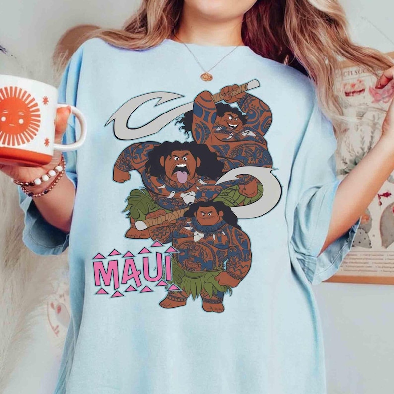 K&ouml;nnte beinhalten: Ein hellblaues T-Shirt mit einem Grafikdruck der Figur Maui aus dem Film Moana in drei verschiedenen Posen, die einen gro&szlig;en Haken halten. Der Text "MAUI" ist in rosa Gro&szlig;buchstaben unter den Figuren mit rosa Dreiecken aufgedruckt.