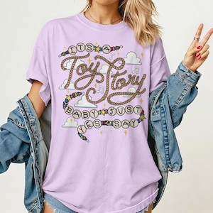 May include: A light purple short-sleeved t-shirt featuring a graphic print with the text "ITS A Toy Story BABY JUST SAY" written in a rope-style font, surrounded by illustrations of clouds, musical notes, stars, and colorful beads.