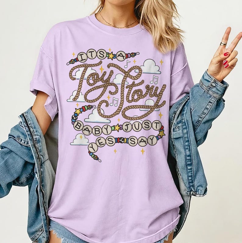 May include: A light purple short-sleeved t-shirt featuring a graphic print with the text "ITS A Toy Story BABY JUST SAY" written in a rope-style font, surrounded by illustrations of clouds, musical notes, stars, and colorful beads.