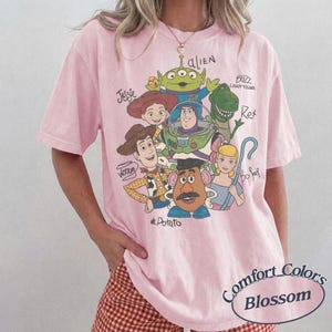 May include: A light pink short-sleeve t-shirt featuring a graphic print of Toy Story characters including Woody, Jessie, Buzz Lightyear, Rex, an alien, Mr. Potato Head, and Bo Peep, with their names written in cursive script around the colorful illustration.