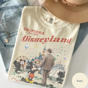 Puede incluir: Camiseta color marfil con un gr&aacute;fico de Disneyland. El dise&ntilde;o presenta a Mickey Mouse, un hombre con traje e im&aacute;genes del parque. El texto dice "CALIFORNIA Est. 1955 Disneyland". La camiseta est&aacute; doblada con un sombrero y vaqueros.