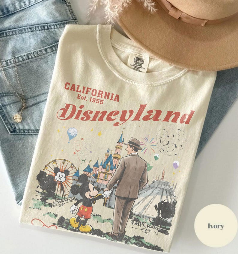 Puede incluir: Camiseta color marfil con un gr&aacute;fico de Disneyland. El dise&ntilde;o presenta a Mickey Mouse, un hombre con traje e im&aacute;genes del parque. El texto dice "CALIFORNIA Est. 1955 Disneyland". La camiseta est&aacute; doblada con un sombrero y vaqueros.
