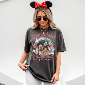 May include: Dark gray t-shirt featuring a Disneyland California graphic with Mickey Mouse, Minnie Mouse, Donald Duck, Goofy, and Pluto. The shirt has a vintage look and feel, with the text "Est 1955". The person is wearing a Minnie Mouse ear headband.