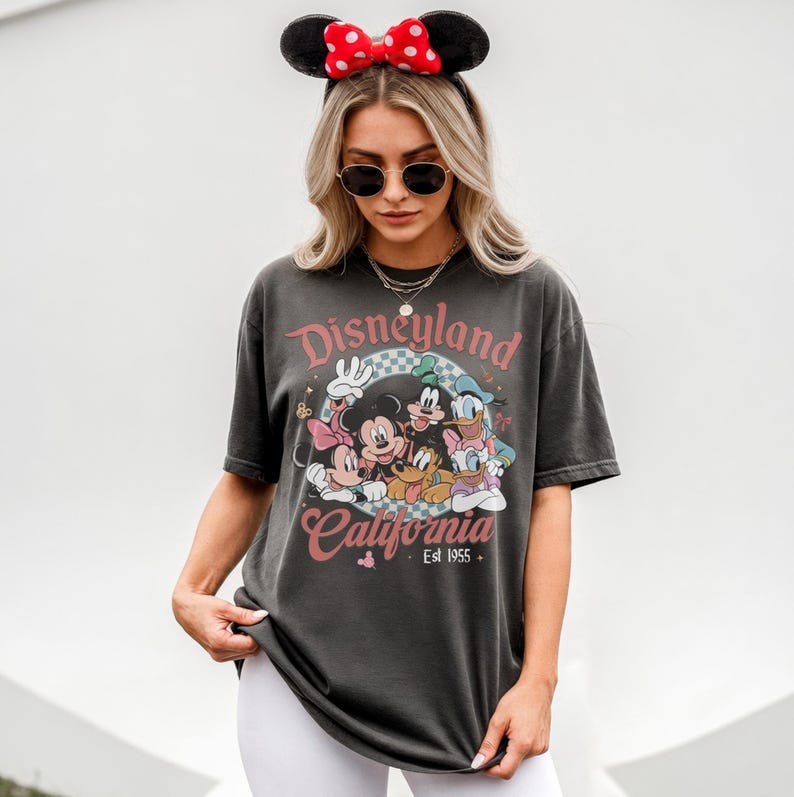 May include: Dark gray t-shirt featuring a Disneyland California graphic with Mickey Mouse, Minnie Mouse, Donald Duck, Goofy, and Pluto. The shirt has a vintage look and feel, with the text "Est 1955". The person is wearing a Minnie Mouse ear headband.