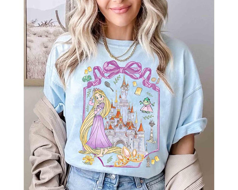 K&ouml;nnte beinhalten: Hellblaues T-Shirt mit einer Cartoon-Illustration einer Prinzessin mit langen blonden Haaren, einem Schloss und anderen M&auml;rchenelementen. Das Design umfasst ein rosa Band, eine Krone und einen Frosch. Das T-Shirt hat hochgekrempelte &Auml;rmel.