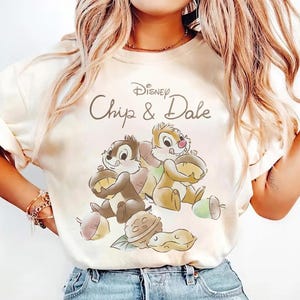 May include: Cream-colored t-shirt featuring the Disney characters Chip and Dale. The graphic shows the two chipmunks holding acorns, with the text "Disney Chip & Dale" above them. The shirt has a tie-dye effect.