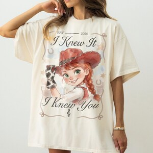 Puede incluir: Una camiseta color crema con el gr&aacute;fico de un personaje de dibujos animados con sombrero de vaquero rojo y guante de estampado de vaca. El texto dice EST 2026, I Knew It e I Knew You, enmarcado por un borde de cuerda con estrellas y herraduras.