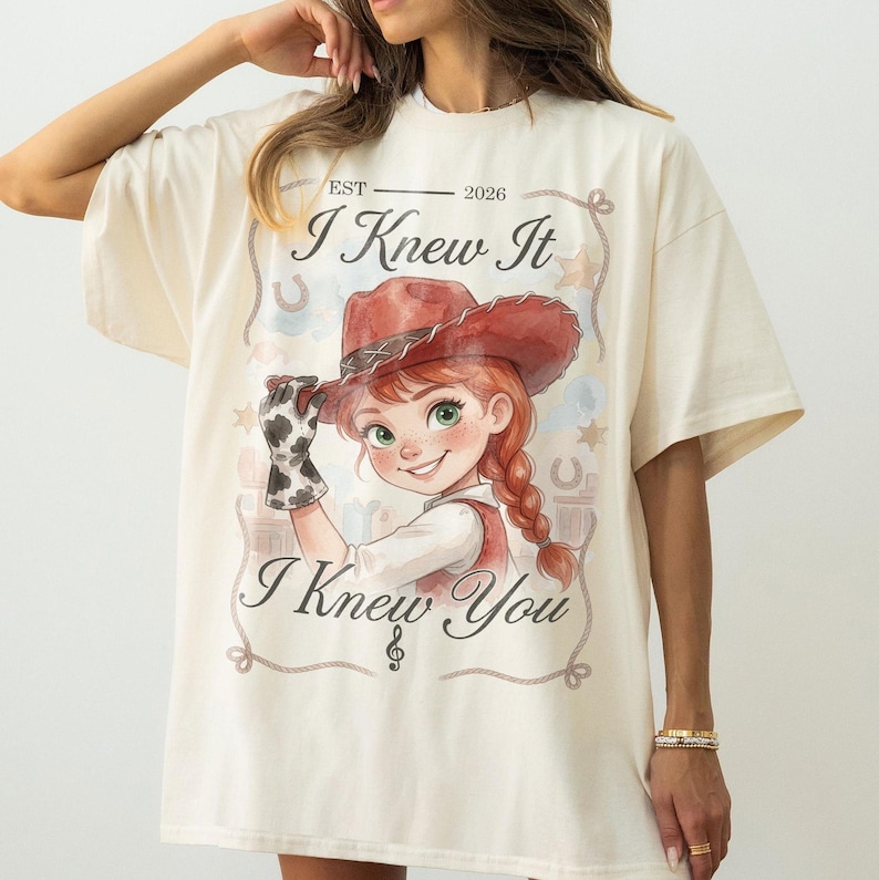 Puede incluir: Una camiseta color crema con el gr&aacute;fico de un personaje de dibujos animados con sombrero de vaquero rojo y guante de estampado de vaca. El texto dice EST 2026, I Knew It e I Knew You, enmarcado por un borde de cuerda con estrellas y herraduras.