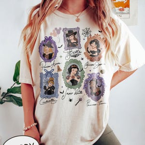 May include: Ivory-colored t-shirt featuring framed portraits of Disney princesses, including Aurora, Belle, Jasmine, Cinderella, Snow White, and Tiana. The shirt has a vintage aesthetic with decorative elements like bats and spiderwebs.