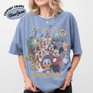 May include: Blue t-shirt with a Disneyland graphic featuring Mickey Mouse, Minnie Mouse, Donald Duck, Goofy, and other characters. The shirt has the text "Disneyland Est. 1955" and "Comfort Colors Blue Jean" on the top left.