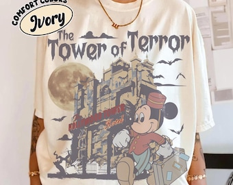 May include: An ivory t-shirt featuring a graphic print of The Tower of Terror with a full moon, bats, and a cartoon mouse character wearing a bellhop uniform and carrying a suitcase. The text reads The Tower of Terror and Hollywood Tower Hotel.