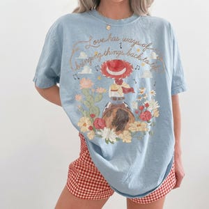 May include: A light blue t-shirt featuring a graphic print of a cowgirl in a red hat riding a horse, surrounded by cacti and flowers. The text reads, Love has ways of bringing things back to life, accompanied by musical notes and small stars.