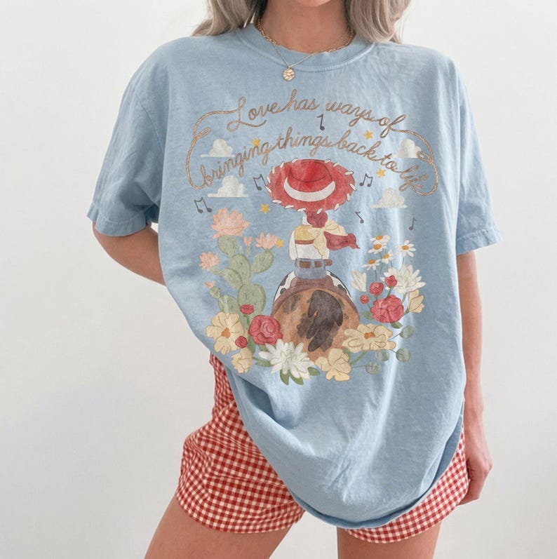 May include: A light blue t-shirt featuring a graphic print of a cowgirl in a red hat riding a horse, surrounded by cacti and flowers. The text reads, Love has ways of bringing things back to life, accompanied by musical notes and small stars.