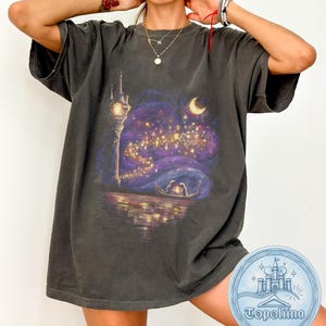 Op de afbeelding: Een antracietgrijs t-shirt met korte mouwen en een grafische print van een hoge stenen toren, een halve maan en een boot met twee figuren op het water, omringd door zwevende lantaarns in een paarse en blauwe nachtelijke hemel.
