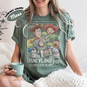 May include: Moss green t-shirt featuring a graphic of Toy Story characters, including Woody, Buzz Lightyear, and Jessie. The design includes the text "DISNEYLAND 2026" and "THE HAPPIEST PLACE ON EARTH."