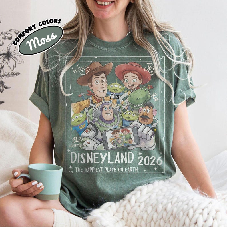 May include: Moss green t-shirt featuring a graphic of Toy Story characters, including Woody, Buzz Lightyear, and Jessie. The design includes the text "DISNEYLAND 2026" and "THE HAPPIEST PLACE ON EARTH."