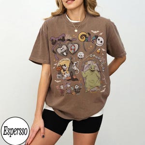 Może przedstawiać: Brązowy t-shirt z kr&oacute;tkim rękawem z nadrukiem graficznym przedstawiającym postacie z Miasteczka Halloween, w tym Jacka Skellingtona, Sally i Oogie Boogie, z napisem NIGHTMARE ułożonym w okrągły wz&oacute;r wok&oacute;ł całego projektu.