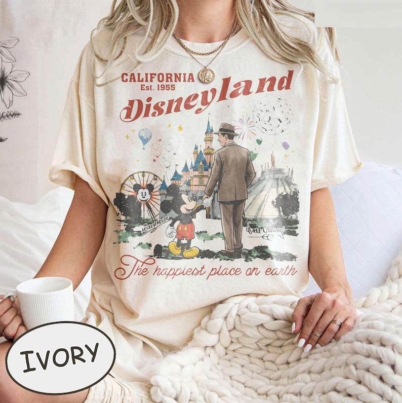 Puede incluir: Camiseta de color marfil con un dise&ntilde;o de Disneyland. El dise&ntilde;o incluye el texto "California Est. 1955 Disneyland" y una ilustraci&oacute;n de Mickey Mouse, un hombre y un castillo. La frase "The happiest place on earth" tambi&eacute;n est&aacute; impresa en la camiseta.