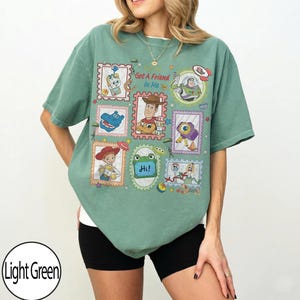 May include: A light green t-shirt featuring a graphic print of various Toy Story characters arranged in postage stamp-style frames. The design includes Woody, Buzz Lightyear, Jessie, and other characters with the text Got A Friend In Me and Hi.