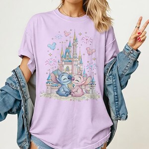 May include: A light purple t-shirt featuring a graphic print of a blue alien character and a pink alien character sitting in front of a large castle. The design includes fireworks, stars, and Mickey-shaped balloons in the background.