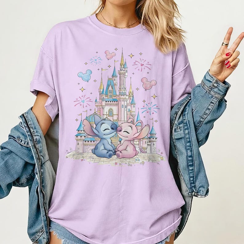 May include: A light purple t-shirt featuring a graphic print of a blue alien character and a pink alien character sitting in front of a large castle. The design includes fireworks, stars, and Mickey-shaped balloons in the background.