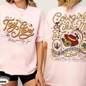 Może przedstawiać: Jasnor&oacute;żowy t-shirt z grafiką z przodu w formie sznurkowego napisu "it's a Toy Story Baby, just say 'yes'" oraz grafiką z tyłu z napisem "Cowgirl like Me", kowbojskim kapeluszem, podkową i napisem "Takes One to Know One".
