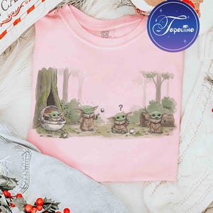 K&ouml;nnte beinhalten: Ein hellrosa T-Shirt mit einer grafischen Illustration von vier kleinen gr&uuml;nen Wesen in einer Waldumgebung. Eines sitzt in einer schwebenden Kapsel, ein anderes greift nach einem Ball, ein drittes wirkt verwirrt mit einem Fragezeichen.