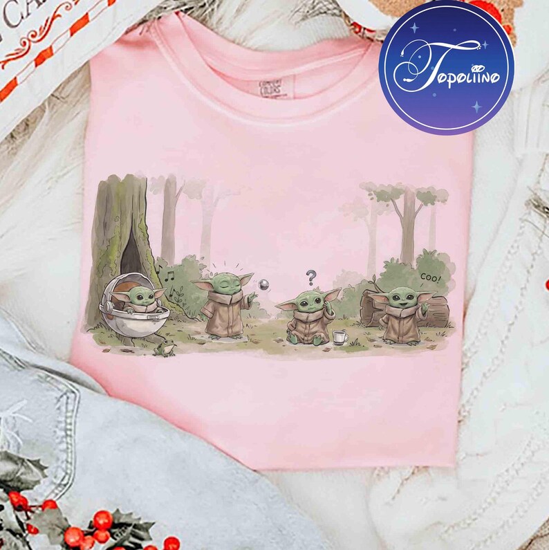 K&ouml;nnte beinhalten: Ein hellrosa T-Shirt mit einer grafischen Illustration von vier kleinen gr&uuml;nen Wesen in einer Waldumgebung. Eines sitzt in einer schwebenden Kapsel, ein anderes greift nach einem Ball, ein drittes wirkt verwirrt mit einem Fragezeichen.