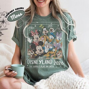 May include: A moss green t-shirt with a vintage Disneyland 2026 graphic featuring Mickey, Minnie, Goofy, Donald, Daisy, Chip, and Dale. The shirt has rolled sleeves and the words "COMFORT COLORS" and "Moss" are printed on the top left.