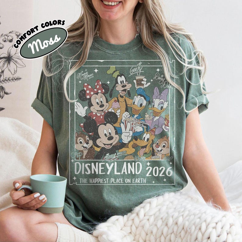 May include: A moss green t-shirt with a vintage Disneyland 2026 graphic featuring Mickey, Minnie, Goofy, Donald, Daisy, Chip, and Dale. The shirt has rolled sleeves and the words "COMFORT COLORS" and "Moss" are printed on the top left.