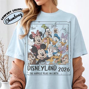 May include: Pale blue t-shirt with a graphic of Disney characters such as Mickey and Minnie Mouse, Donald Duck, and Goofy. The graphic says "DISNEYLAND 2026" and "THE HAPPIEST PLACE ON EARTH". The shirt has the words "COMFORT COLORS Chambray" on the upper left.