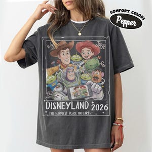 May include: Dark gray t-shirt featuring a graphic of Disney characters including Woody, Buzz Lightyear, and Jessie. The design includes the text "DISNEYLAND 2026" and "THE HAPPIEST PLACE ON EARTH". The shirt has a relaxed fit and the words "COMFORT COLORS" and "Pepper" are printed on the top right.