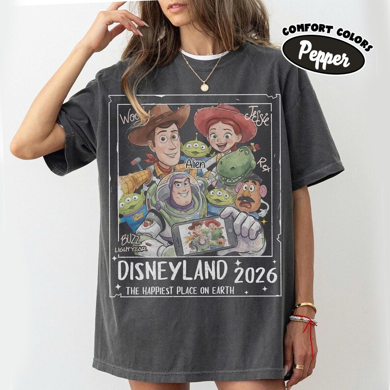May include: Dark gray t-shirt featuring a graphic of Disney characters including Woody, Buzz Lightyear, and Jessie. The design includes the text "DISNEYLAND 2026" and "THE HAPPIEST PLACE ON EARTH". The shirt has a relaxed fit and the words "COMFORT COLORS" and "Pepper" are printed on the top right.