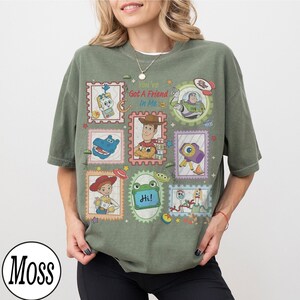 May include: An olive green t-shirt featuring a graphic print of various Toy Story characters arranged in postage stamp-style frames. The text "You've Got A Friend In Me" is printed in red and blue above the central image of Woody the cowboy.