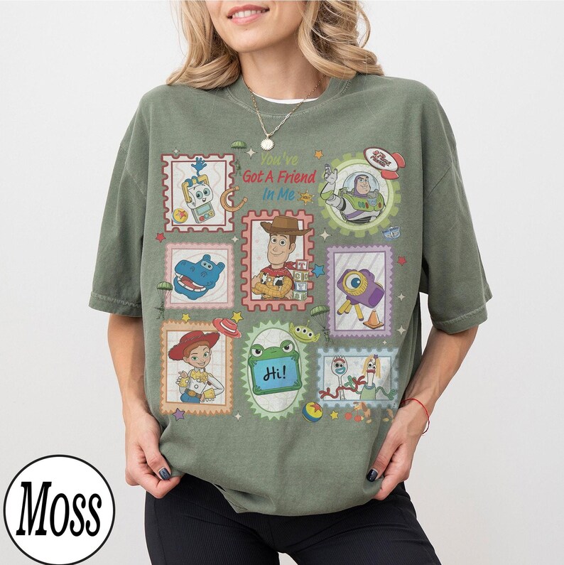 May include: An olive green t-shirt featuring a graphic print of various Toy Story characters arranged in postage stamp-style frames. The text "You've Got A Friend In Me" is printed in red and blue above the central image of Woody the cowboy.