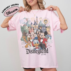 May include: A light pink t-shirt featuring a Disneyland graphic with Mickey Mouse, Minnie Mouse, Donald Duck, Goofy, and Pluto. The shirt has the text "Disneyland Est. 1955" and "Comfort Colors Blossom" on the top left.