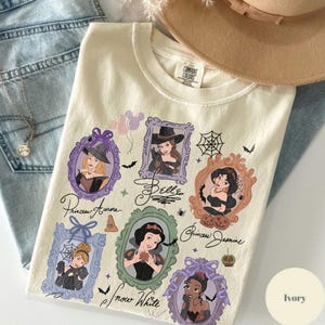 May include: Ivory-colored Comfort Colors t-shirt featuring framed illustrations of Disney princesses in Halloween costumes. The shirt includes the names of the princesses and Halloween-themed graphics like bats and spiderwebs.