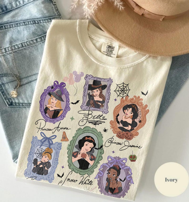 May include: Ivory-colored Comfort Colors t-shirt featuring framed illustrations of Disney princesses in Halloween costumes. The shirt includes the names of the princesses and Halloween-themed graphics like bats and spiderwebs.