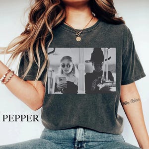 May include: Dark gray t-shirt with a black and white graphic print. The graphic features two people, one wearing sunglasses and holding a phone, and the other wearing a hat. The word "PEPPER" is printed at the bottom of the shirt.