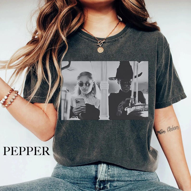 May include: Dark gray t-shirt with a black and white graphic print. The graphic features two people, one wearing sunglasses and holding a phone, and the other wearing a hat. The word "PEPPER" is printed at the bottom of the shirt.