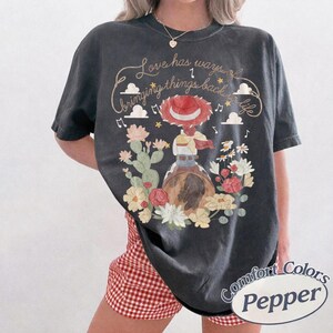May include: A charcoal gray t-shirt featuring a graphic of a cowgirl in a red hat riding a horse, surrounded by cacti and flowers. The text reads, Love has ways of bringing things back to life, accompanied by musical notes and cloud illustrations.