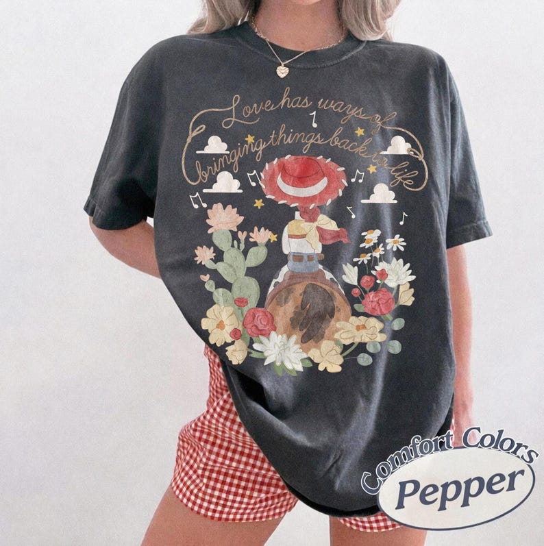 May include: A charcoal gray t-shirt featuring a graphic of a cowgirl in a red hat riding a horse, surrounded by cacti and flowers. The text reads, Love has ways of bringing things back to life, accompanied by musical notes and cloud illustrations.