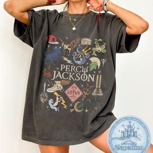 Puede incluir: Una camiseta de manga corta gris carbón con un estampado gráfico que incluye el texto PERCY JACKSON y LOTUS HOTEL. El diseño presenta varias ilustraciones como un caballo alado, un tridente, una espada, un rayo y un rascacielos.