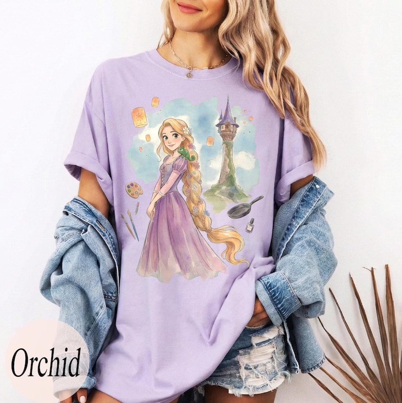 K&ouml;nnte beinhalten: Ein helllila T-Shirt mit einer Aquarellillustration einer Prinzessin mit langen blonden Haaren, einem Turm und schwebenden Laternen. Das Wort "Orchid" ist unten links auf dem T-Shirt aufgedruckt.