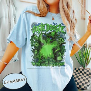 Op de afbeelding: Lichtblauw T-shirt met een groene afbeelding van het Oogie Boogie-personage uit The Nightmare Before Christmas. De afbeelding bevat de tekst "Oogie Boogie" en "I'm the Oogie Boogie Man!" in het groen. Het shirt is chambray van kleur.
