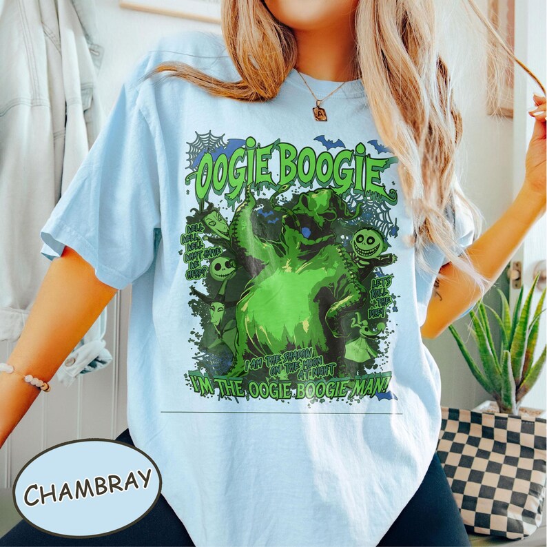 Op de afbeelding: Lichtblauw T-shirt met een groene afbeelding van het Oogie Boogie-personage uit The Nightmare Before Christmas. De afbeelding bevat de tekst "Oogie Boogie" en "I'm the Oogie Boogie Man!" in het groen. Het shirt is chambray van kleur.