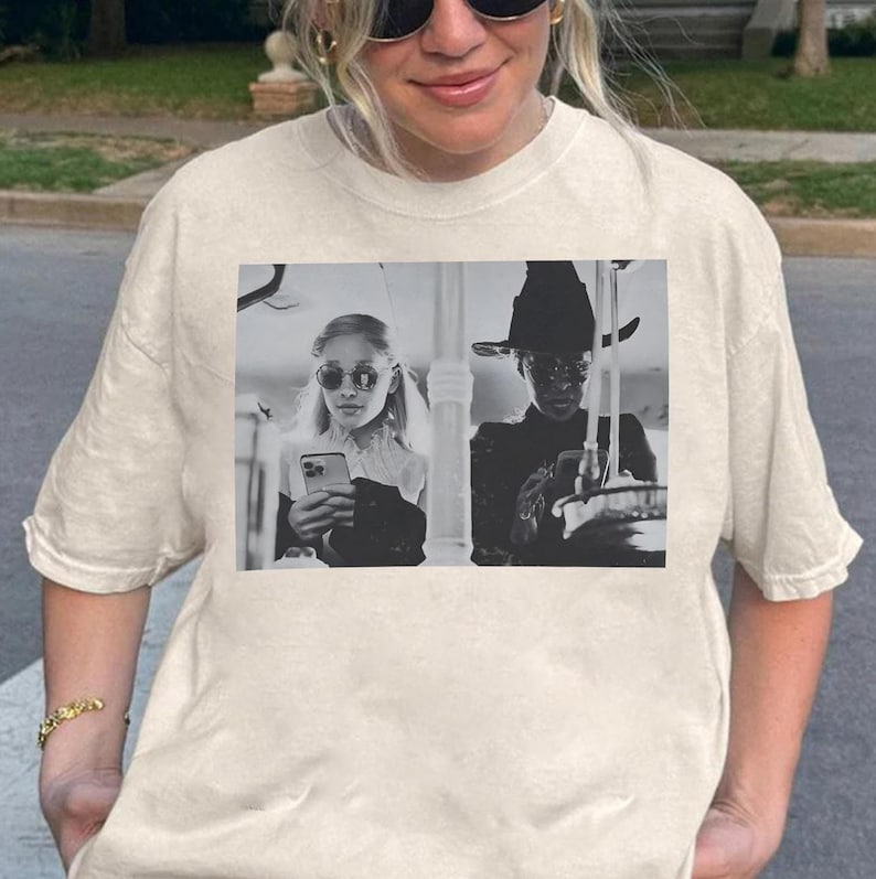 May include: Cream-colored t-shirt featuring a black and white image of two people, one wearing a witch hat and the other wearing sunglasses, both holding phones. The t-shirt is a casual, everyday wear item.