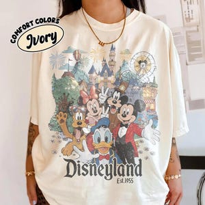 May include: Ivory-colored t-shirt featuring a Disneyland graphic with Mickey Mouse, Minnie Mouse, Donald Duck, and other characters. The graphic includes a castle, fireworks, and the text "Disneyland Est. 1955."