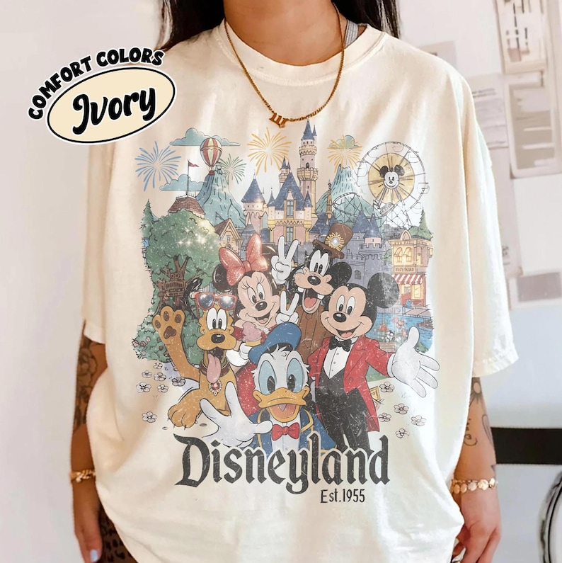 May include: Ivory-colored t-shirt featuring a Disneyland graphic with Mickey Mouse, Minnie Mouse, Donald Duck, and other characters. The graphic includes a castle, fireworks, and the text "Disneyland Est. 1955."